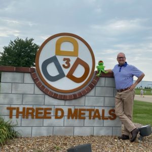 Male Three D Metals employee standing next to the Three D Metals sign with a stuffed Kermit the Frog.