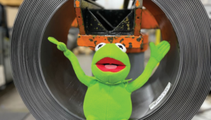 Stuffed Kermit the Frog sitting on a roll of high carbon steel on the shop floor.