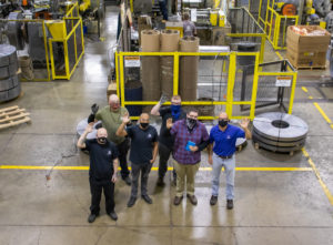 Group of Three D Metals employees standing on the shop floor and waving while wearing masks.